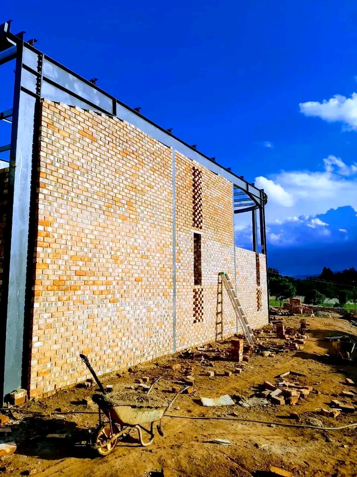 Warehouse construction with brick walls and steel roof framework, wheelbarrow on muddy construction site