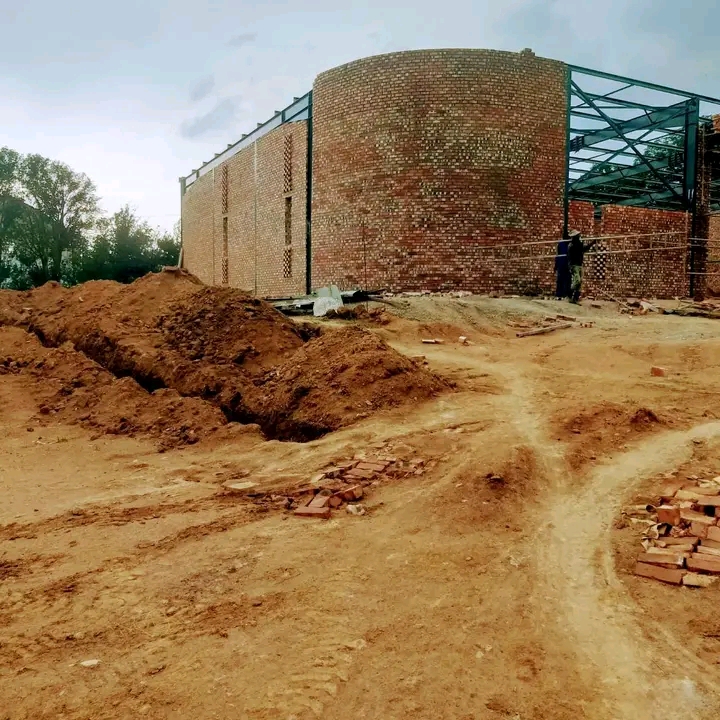 Large-scale warehouse construction with curved brick walls and steel truss framework, extensive earthworks